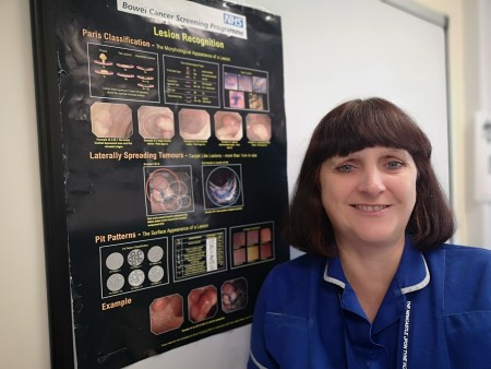 A woman with short, dark hair standing next to a poster for the Bowel Cancer Screening Programme.