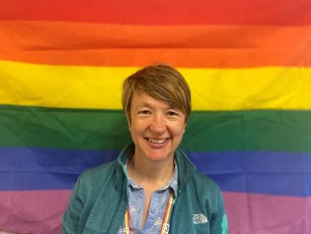 A woman with short brown hair standing in front of a rainbow flag.