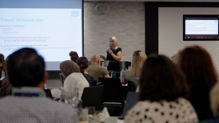 Woman with blonde hair wearing a black dress speaking at a conference in front of a crowd of people 